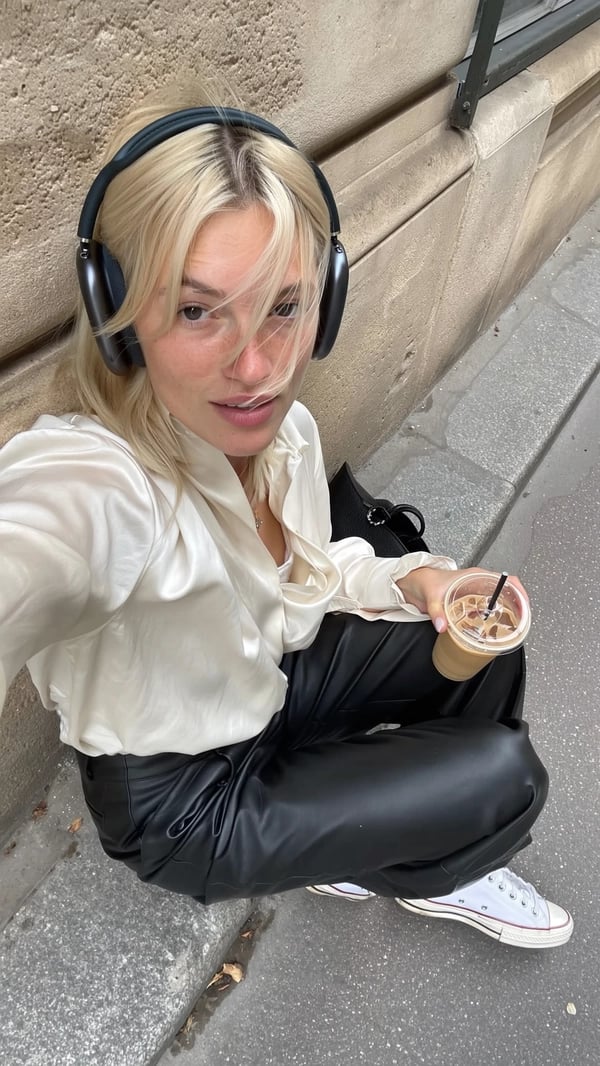 A spontaneously captured iPhone-styled candid photo of a young woman with platinum hair casually lounging against a textured, slightly weathered Parisian stone wall on a city sidewalk. She wears understated effortless cool conveyed through an ivory silk blouse from COS with soft draping and subtle fabric wrinkles paired with sleek black leather pants by Our Legacy, complemented by classic white Converse sneakers gently scuffed from wear. Over her hair sit minimal black headphones, resting lightly and adding a contemporary casual touch. In one hand, she holds a translucent iced coffee glass showing realistic condensation and subtle reflections. Her hair is styled in a naturally tousled manner, some strands catching soft natural daylight that gently illuminates her neutral makeup and natural skin texture, including faint freckles and fine pores. Surrounding her are authentic urban textures of worn concrete pavement and the rough stone wall with patches of subtle moss. The composition features a slightly tilted overhead angle, off-center framing, and an informal crop that evoke the intuitive spontaneity and genuine intimacy emblematic of everyday Parisian iPhone street photography. Soft shadows and delicate highlights reveal the delicate fabric fibers, realistic leather grain, and glass transparency, completing the true-to-life candid aesthetic.